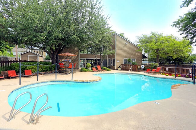 Outdoor swimming pool with curved edges, red lounge chairs, and shaded patio area surrounded by trees and greenery - Brittany Square Apartments