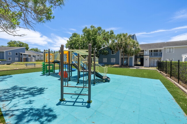 Charming community playground with vibrant equipment surrounded by lush greenery and modern apartment buildings under a clear blue sky. - Vista North